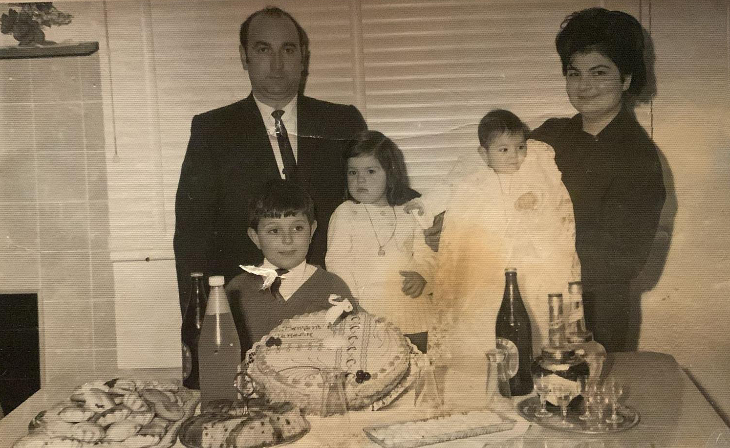 Questa foto, datata 1970, ritrae una famiglia conflentese emigrata Melbourne in Australia e inviata a Conflenti, paese calabrese in provincia di Catanzaro, accompagnata da un messaggio di auguri. La famiglia, composta da moglie, marito e e tre bambini al centro, è in posa dietro un tavolo imbandito a festa con una torta, pasticcini, bibite e bottiglie di liquori, probabilmente in occasione del compleanno di uno dei bambini.